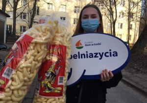 Uczennica stoi i trzyma w ręku chrupki kukurydziane, dwie duże paczki.